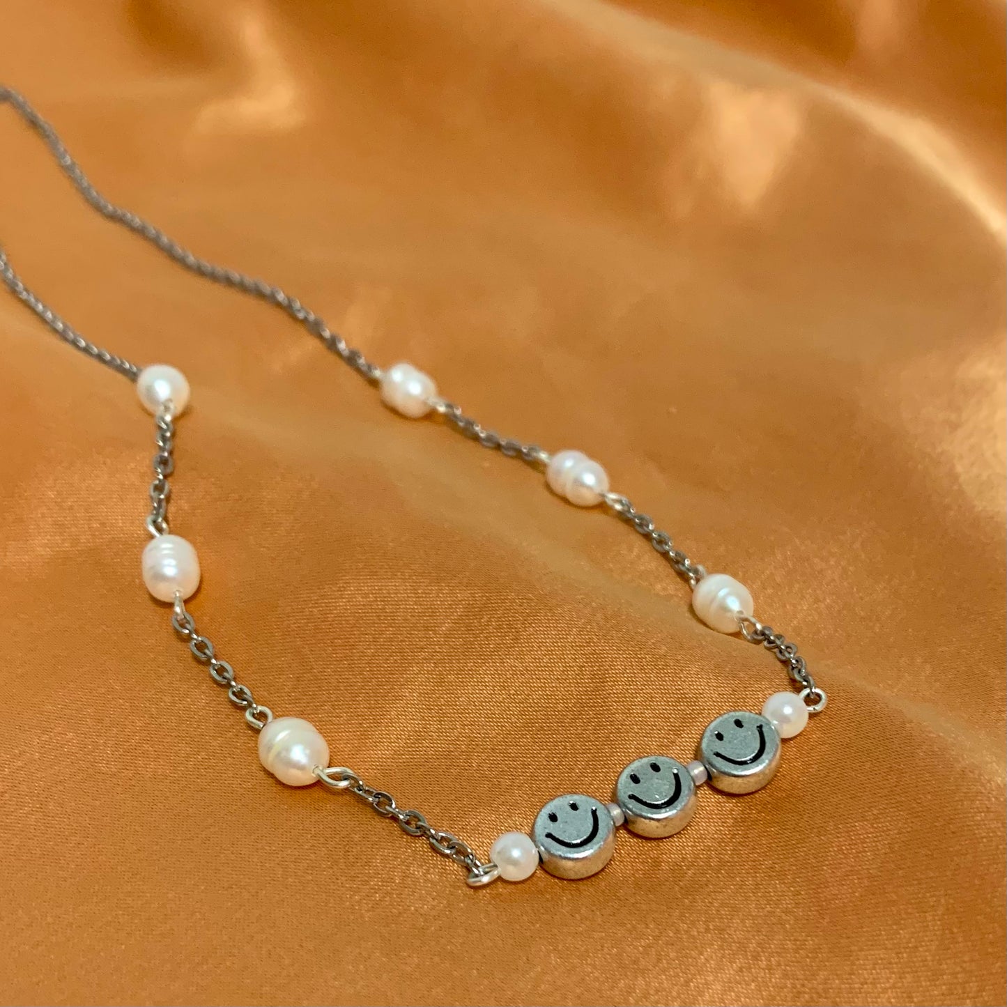 Silver chain necklace connected by freshwater pearls and a wire bar featuring three silver smiley face beads and pearls between each on an orange satin background.
