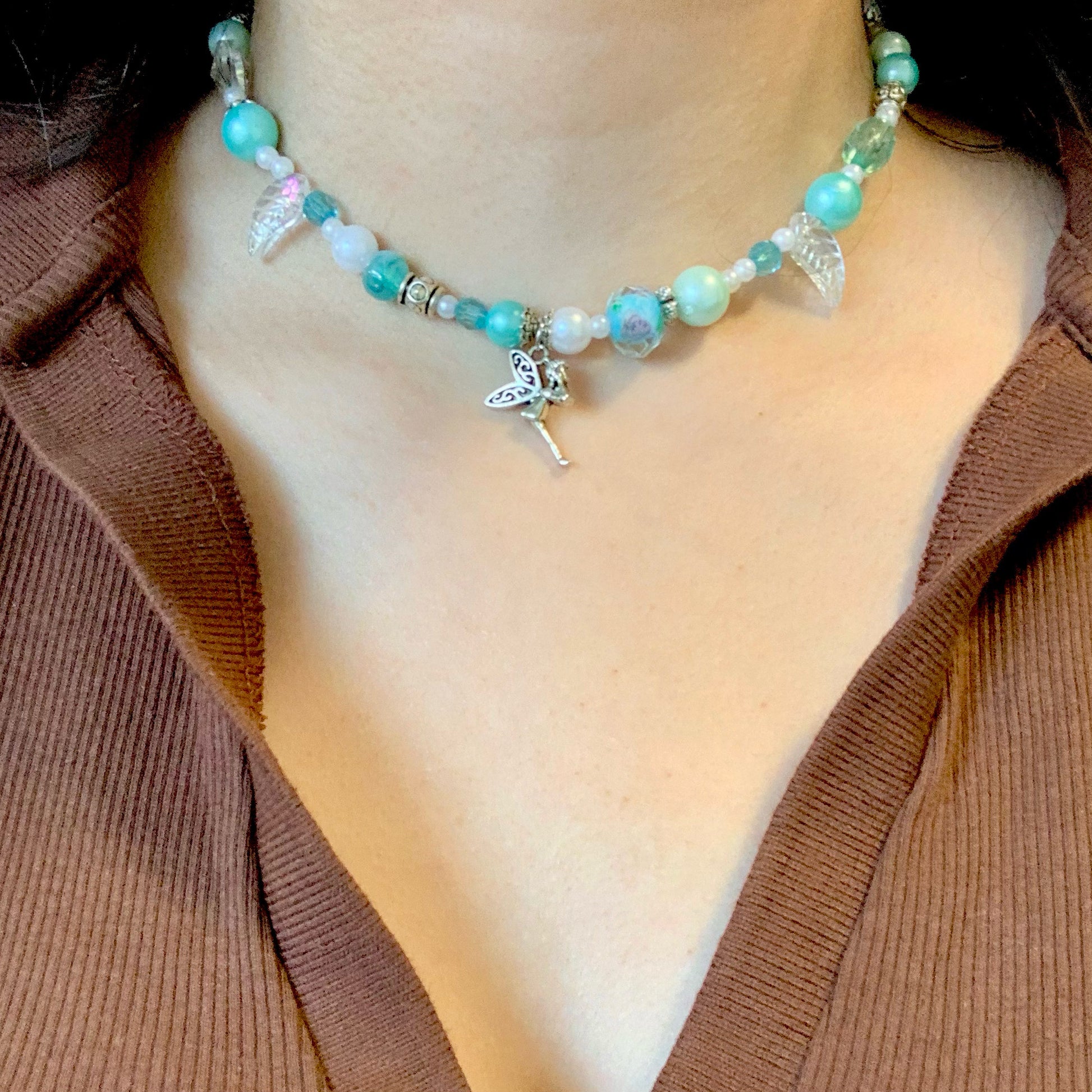 Wearing a turquoise and white glass bead necklace with a fairy charm and a brown shirt.