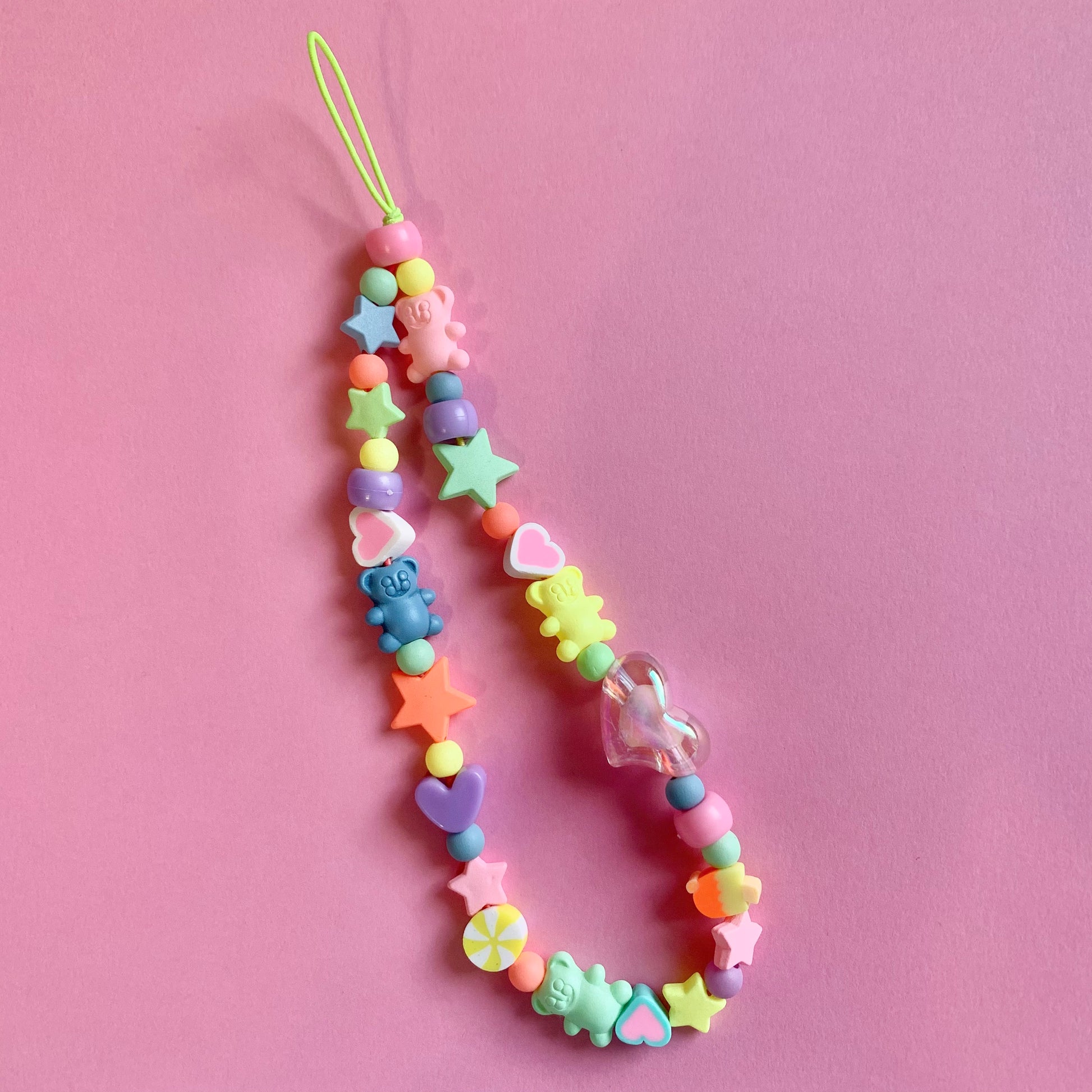 Pastel phone charm featuring teddy bear beads, stars, and a statement heart bead. Photographed on a pink background.